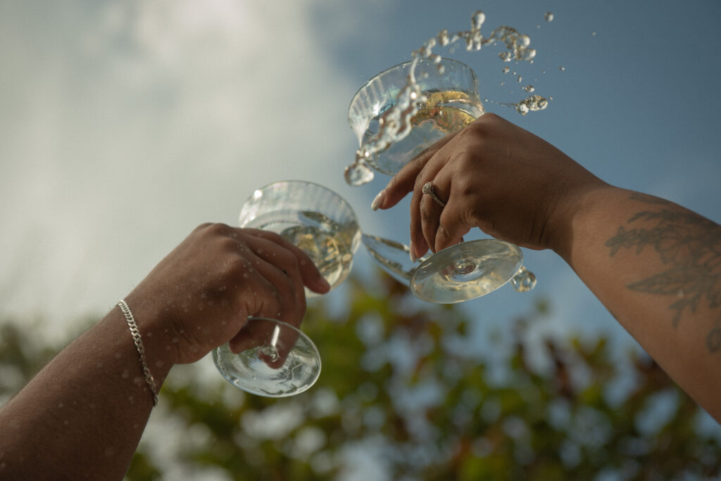Hands holding coupe glasses in mid-toast as champagne splashes in the sunlight during a garden engagement celebration.
