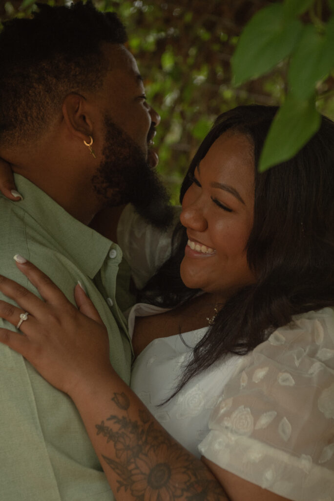 A couple embraces closely under lush greenery, sharing a warm and candid moment during their botanical garden engagement session.