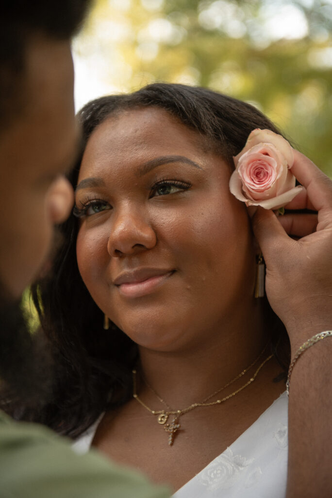 A close-up engagement portrait of a woman smiling softly as a rose is tucked behind her ear at Mercer Botanical Gardens.
