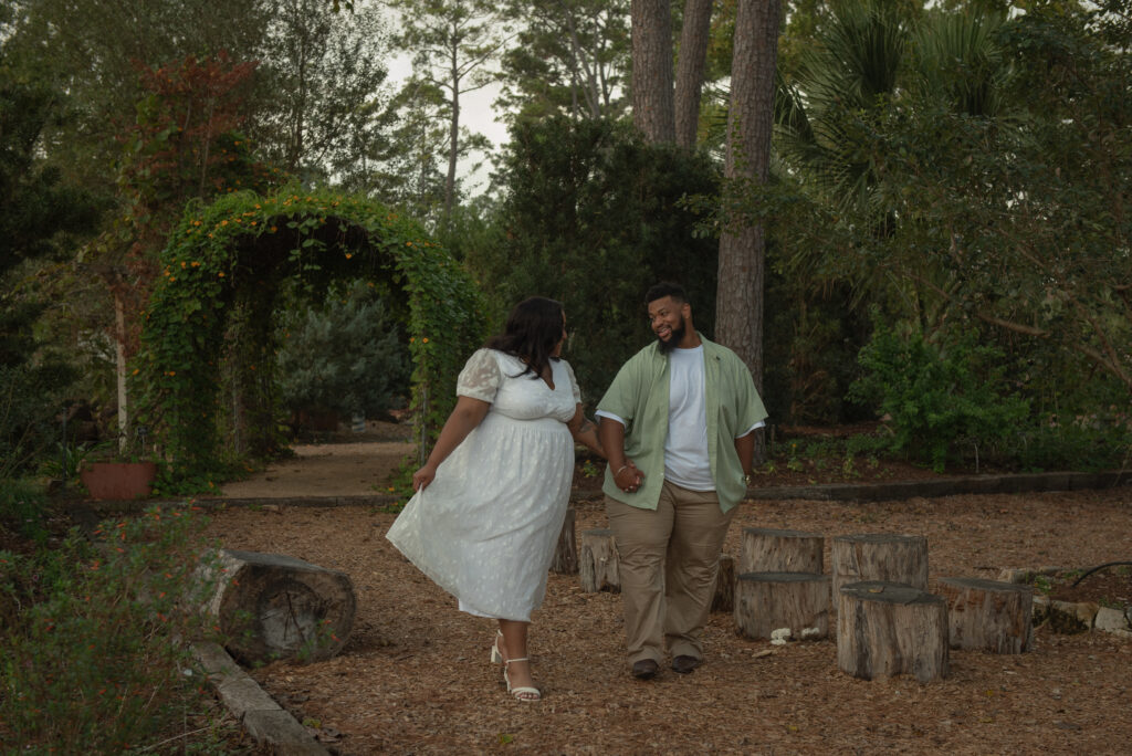 A couple walks hand-in-hand through a rose garden pathway during a romantic Houston engagement session.