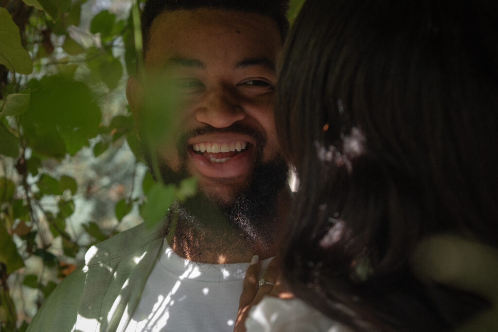 A candid engagement photo of a man smiling through the leaves, taken during a natural, documentary-style session at Mercer Botanical Gardens