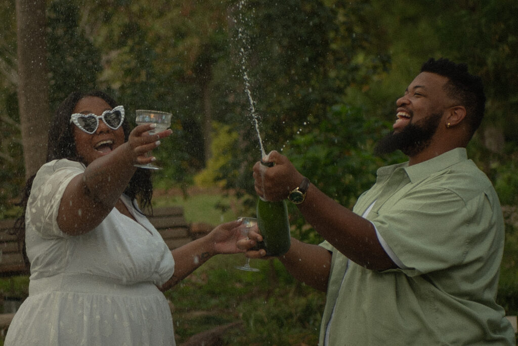 A couple laughs and sprays champagne during a fun, celebratory engagement session at Mercer Botanical Gardens.