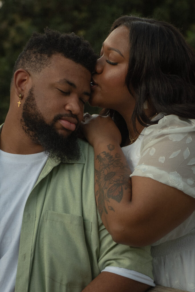 A woman gently kisses her partner’s forehead in a quiet, intimate moment during a romantic garden engagement session.