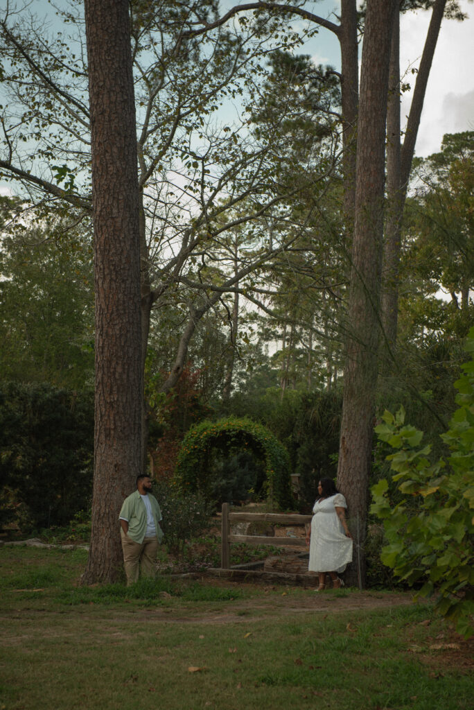 A couple stands facing one another between tall trees in a quiet forested area of Mercer Botanical Gardens.