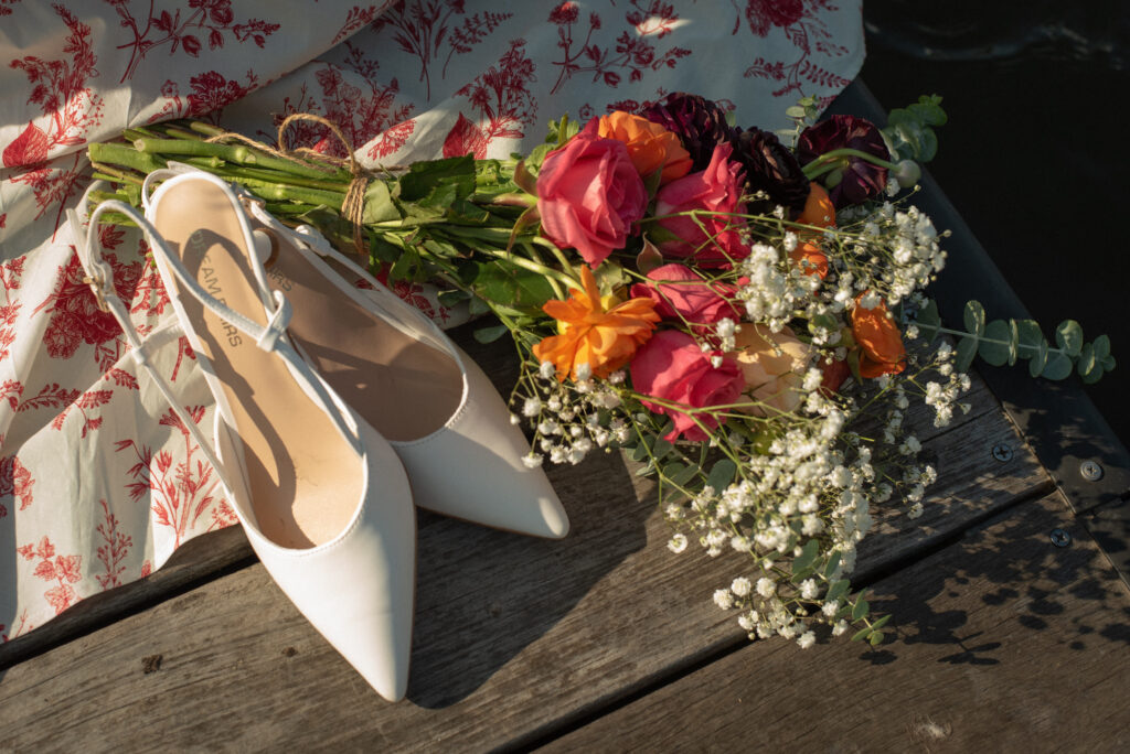 Editorial detail shot of white heels and a colorful bouquet resting on a floral dress during a romantic lakeside proposal at Lake Woodlands.
