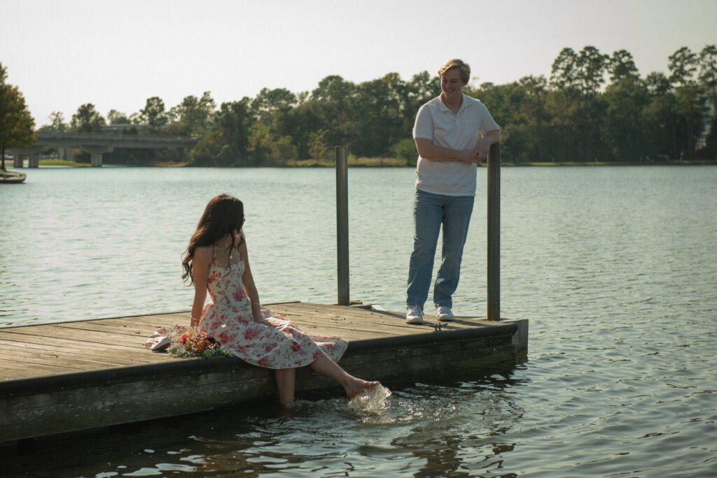 Romantic lakeside moment with woman sitting on dock splashing her feet while her partner watches nearby.