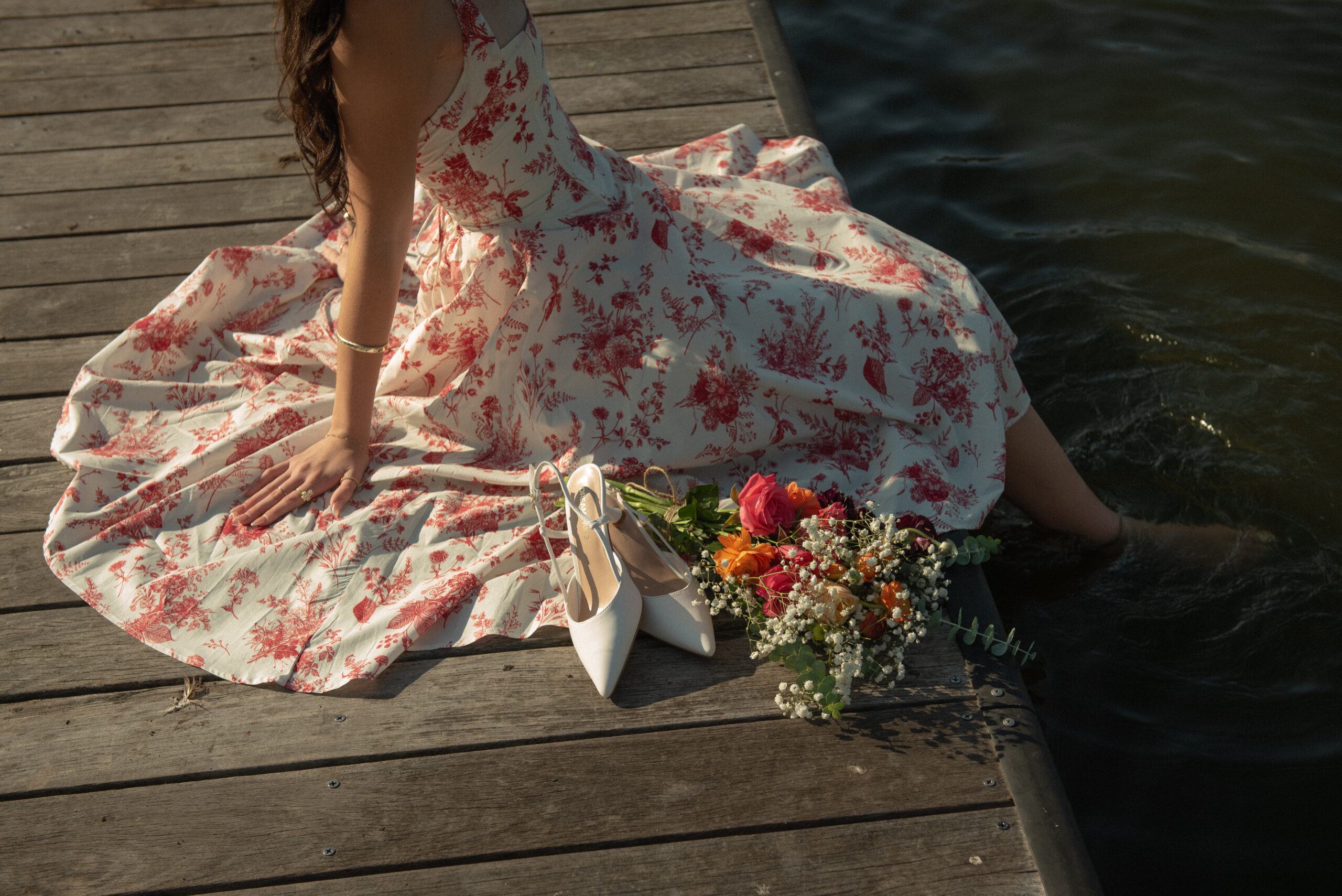 Editorial detail shot of floral dress, white heels, and bouquet on dock during a lakeside proposal session
