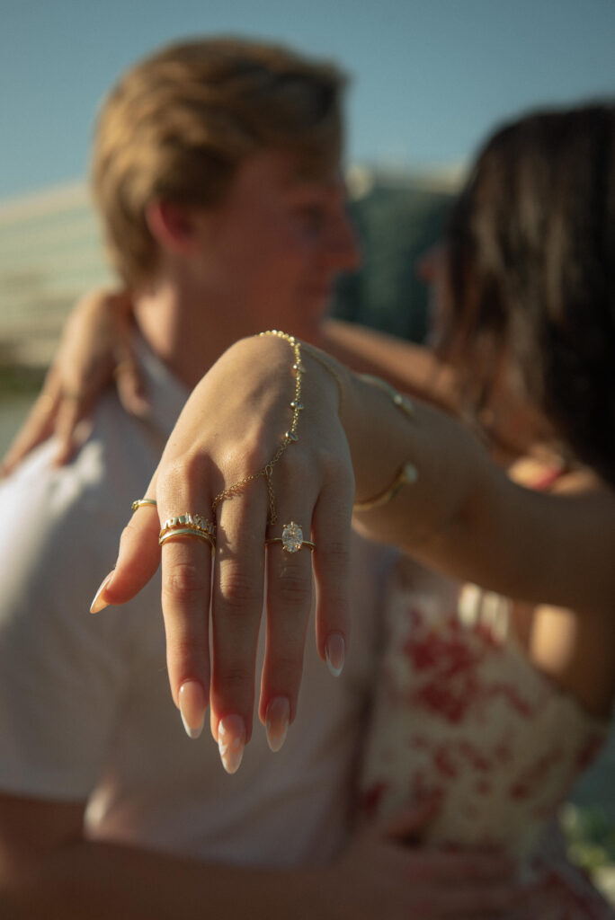 Editorial engagement ring close-up with couple embracing in the background during lakeside proposal session.