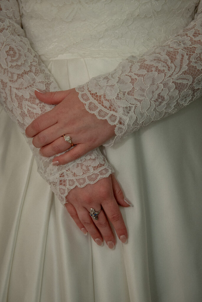 A close-up of the bride’s lace sleeves and wedding rings as she gently rests her hands together.