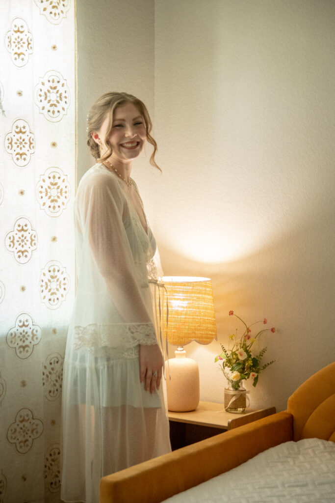 The bride stands near a window in soft natural light as she gets ready before the ceremony.