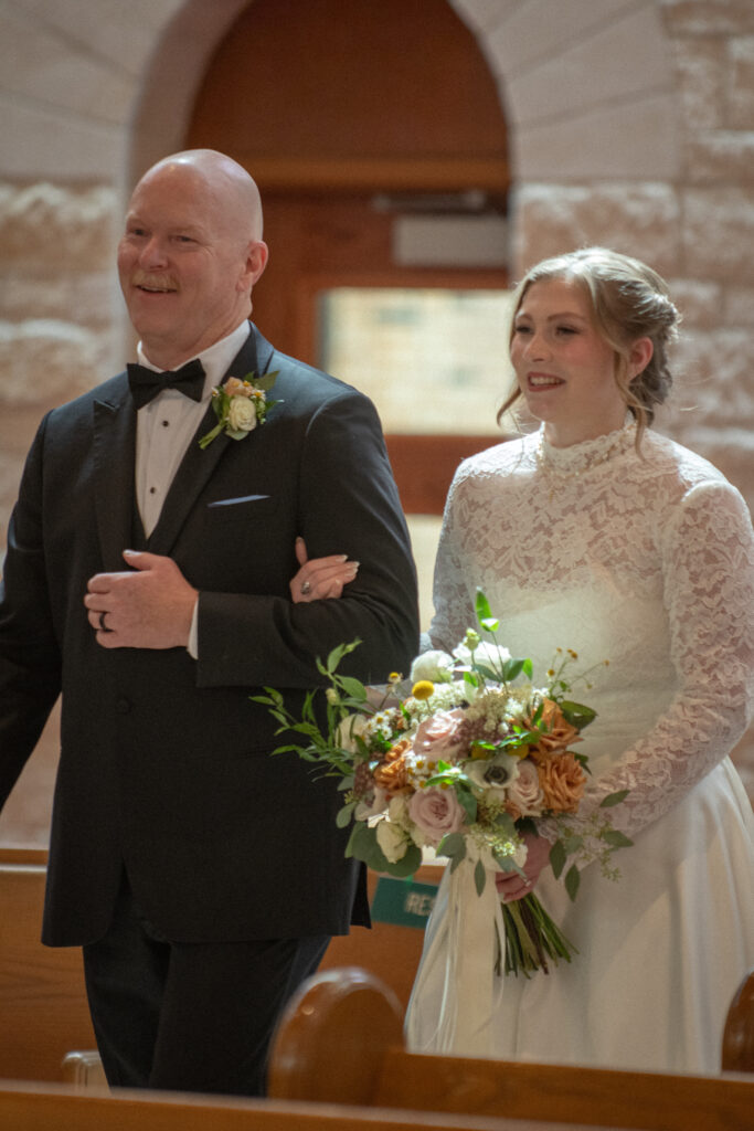 A bride walks down the aisle with her father, holding a bouquet before the wedding ceremony begins.