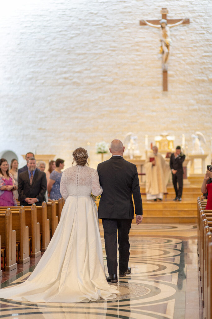 A bride and her father walk arm-in-arm into the church toward the altar at the start of the wedding ceremony.