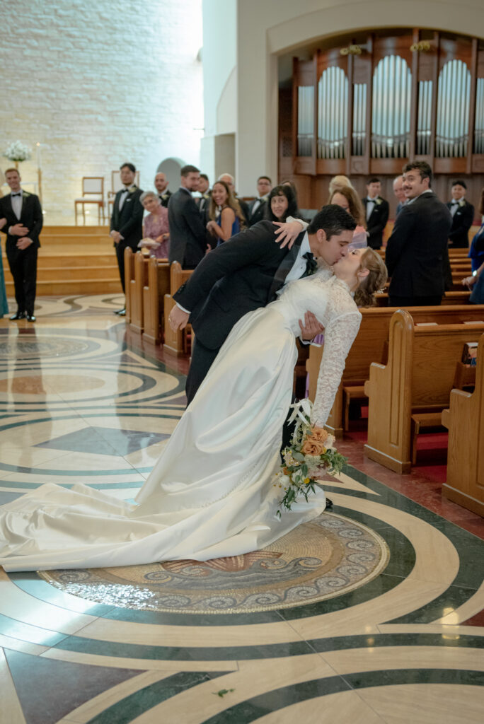 Bride and groom share a dip kiss down the aisle at St. John Vianney Catholic Church in Houston as guests cheer.