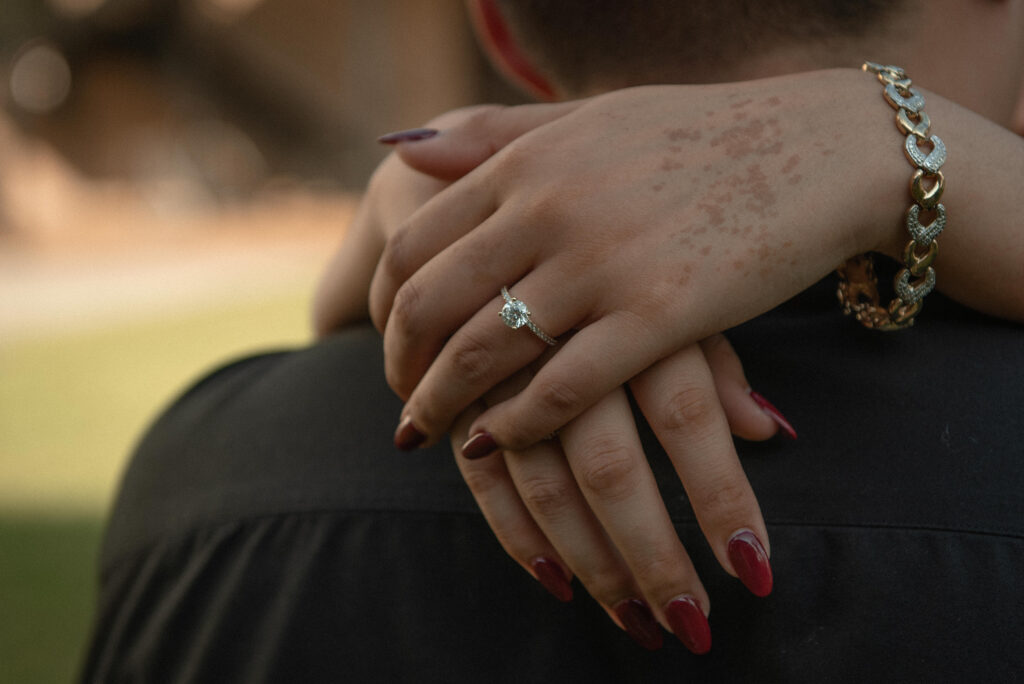 A close-up of a woman’s hands wrapped around her partner, showing her engagement ring.