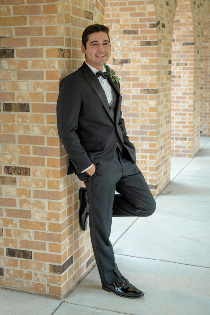 A groom leans casually against a brick wall while wearing a classic black tuxedo before the wedding.