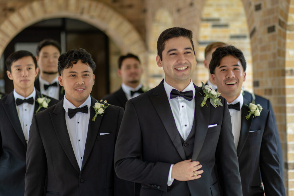 A groom and his groomsmen walk together in matching black tuxedos before the wedding ceremony.