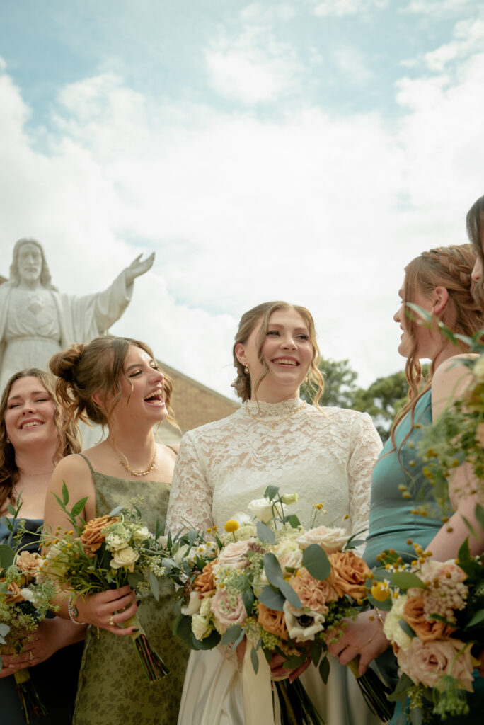 Bride smiles with her bridesmaids holding floral bouquets outside St. John Vianney Catholic Church.