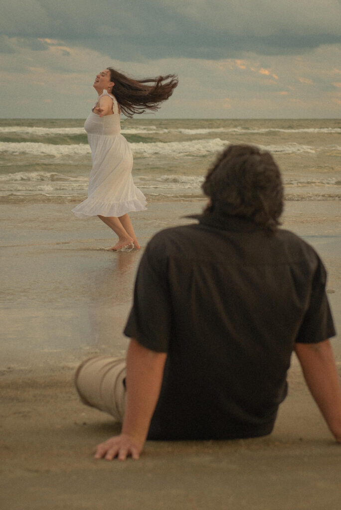 Woman twirling in the water while her partner watches during a Galveston engagement session.