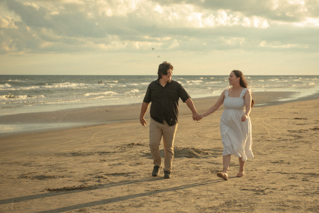 Couple smiling as they walk toward the camera along the shoreline at sunset.