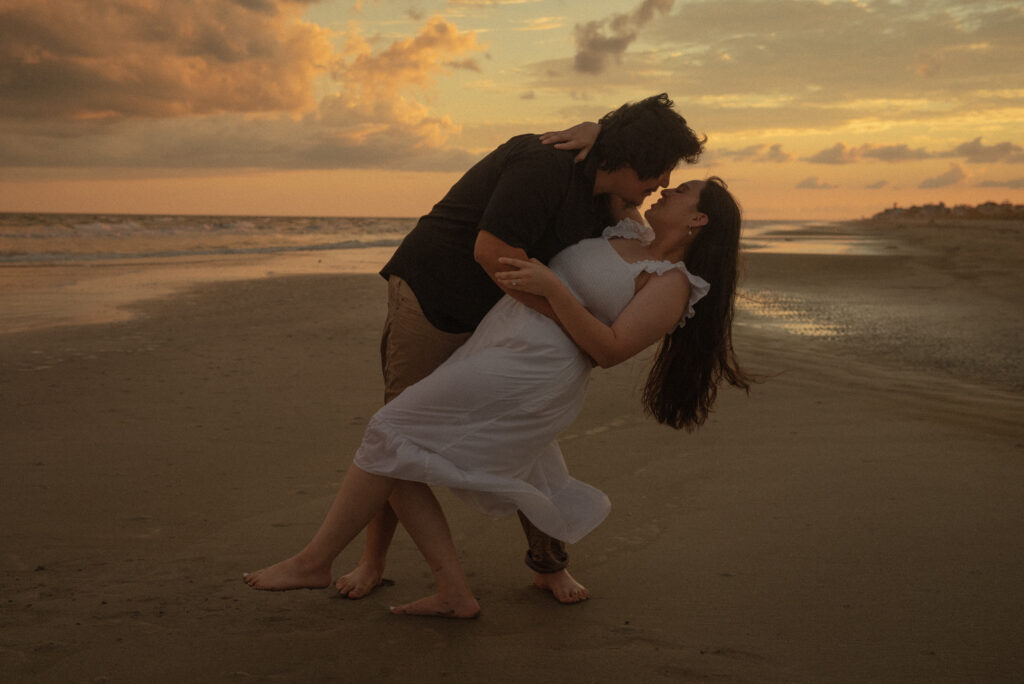 Couple sharing a dip kiss on the beach during a warm sunset engagement session.
