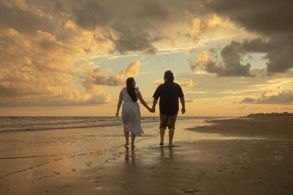 Engaged couple holding hands and walking away from the camera during a golden sunset.