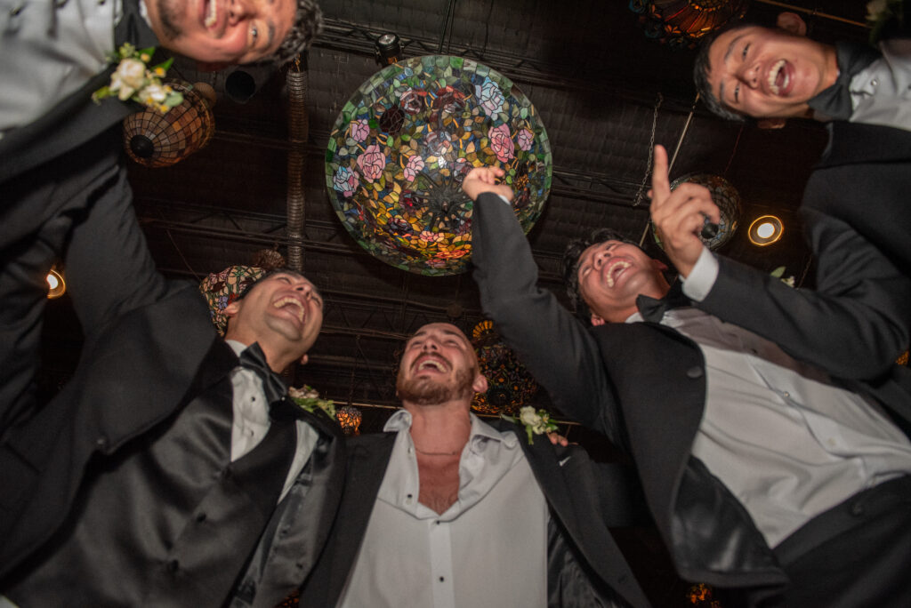 Groomsmen cheer and dance beneath the stained-glass lamps at the reception.