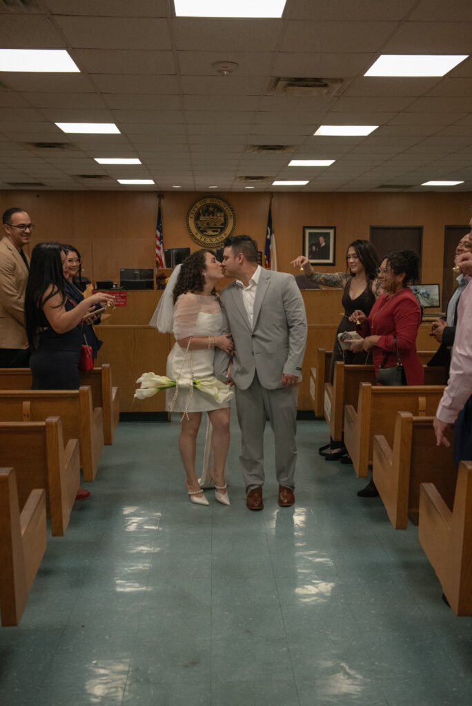 Couple sharing a just married kiss in a Houston municipal courthouse courtroom with family cheering nearby.