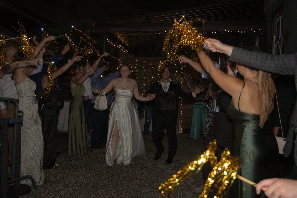 Bride and groom walk through a tunnel of guests waving gold tinsel wands during their wedding exit.