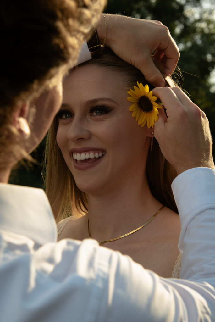Summer engagement photo of a woman smiling as her partner places a sunflower behind her ear during golden hour