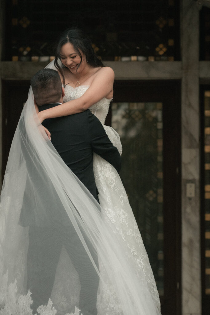 Bride laughing while being lifted by the groom, veil flowing behind them, joyful romantic wedding portrait.