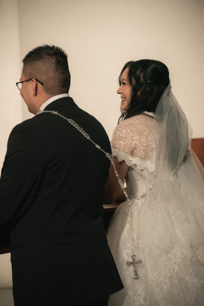 Couple smiling together in church pews during a Catholic wedding ceremony.