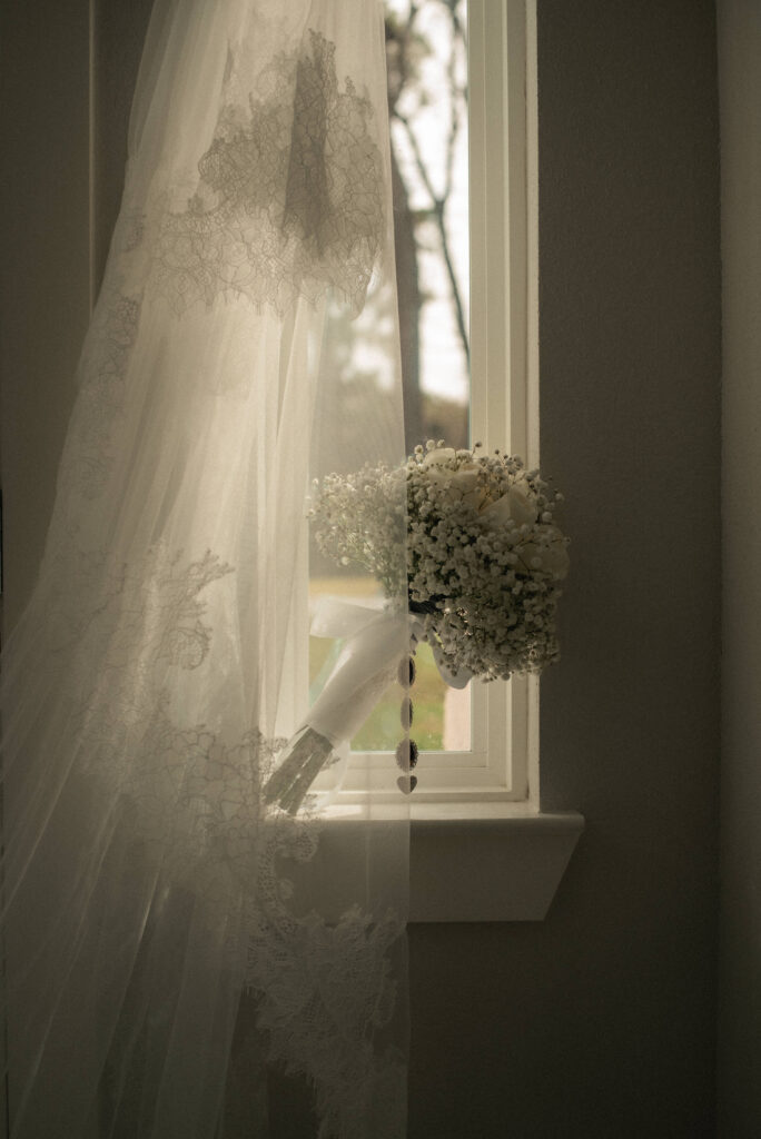 Bridal veil hanging beside a window with a wedding bouquet resting in soft natural light.