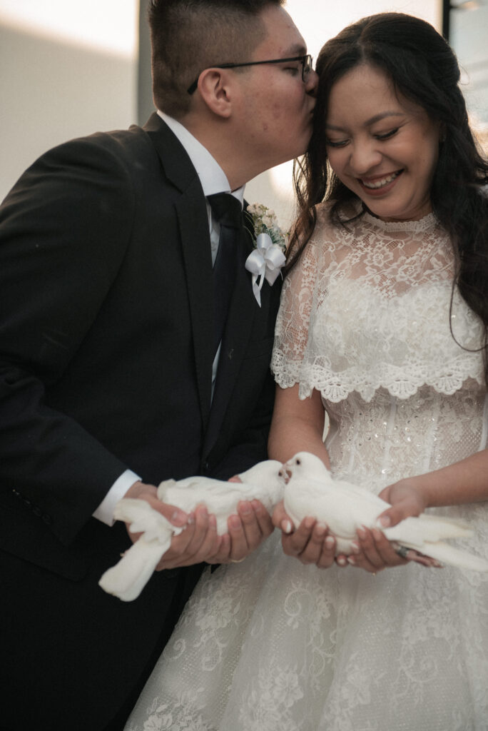 Newlyweds holding white doves as the groom kisses the bride on the forehead, romantic wedding moment outdoors.