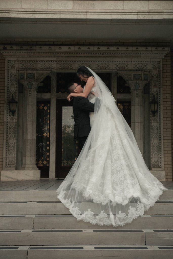 Groom lifting bride in front of an ornate building entrance, full length romantic wedding portrait with veil and lace gown.