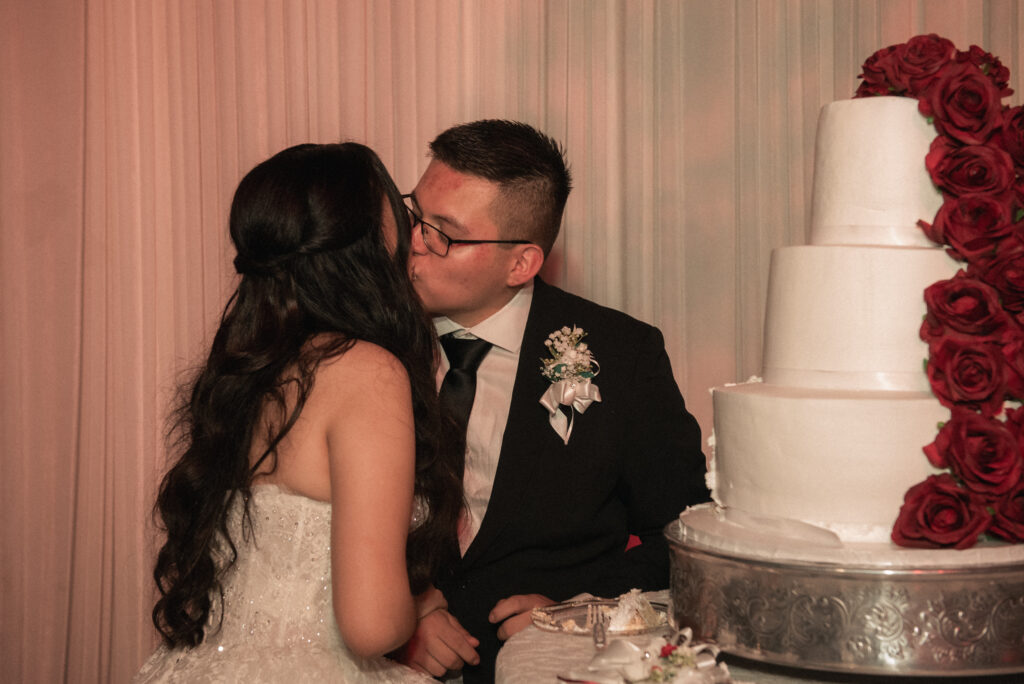 Couple kissing beside a tiered wedding cake decorated with red roses.