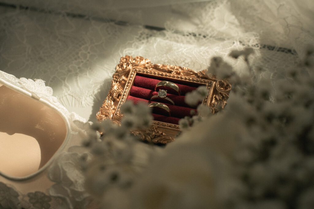 Wedding rings in a vintage gold ring box on lace fabric with soft florals and bridal details.