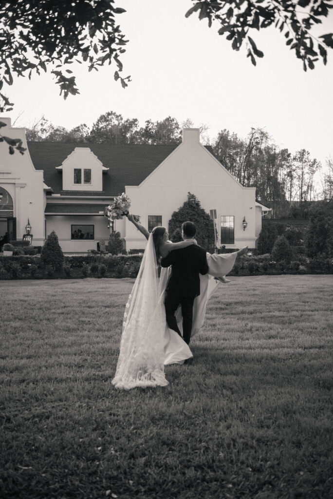 Black and white photo of the groom carrying the bride across a lawn toward the venue.