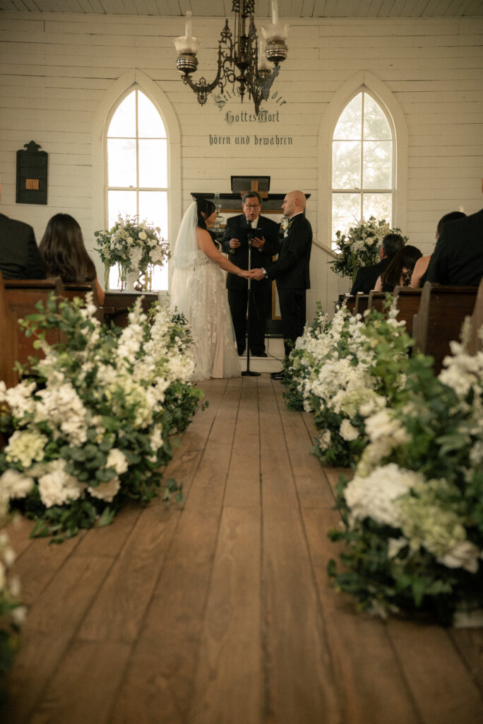 Wide shot of chapel wedding ceremony with floral aisle arrangements and guests