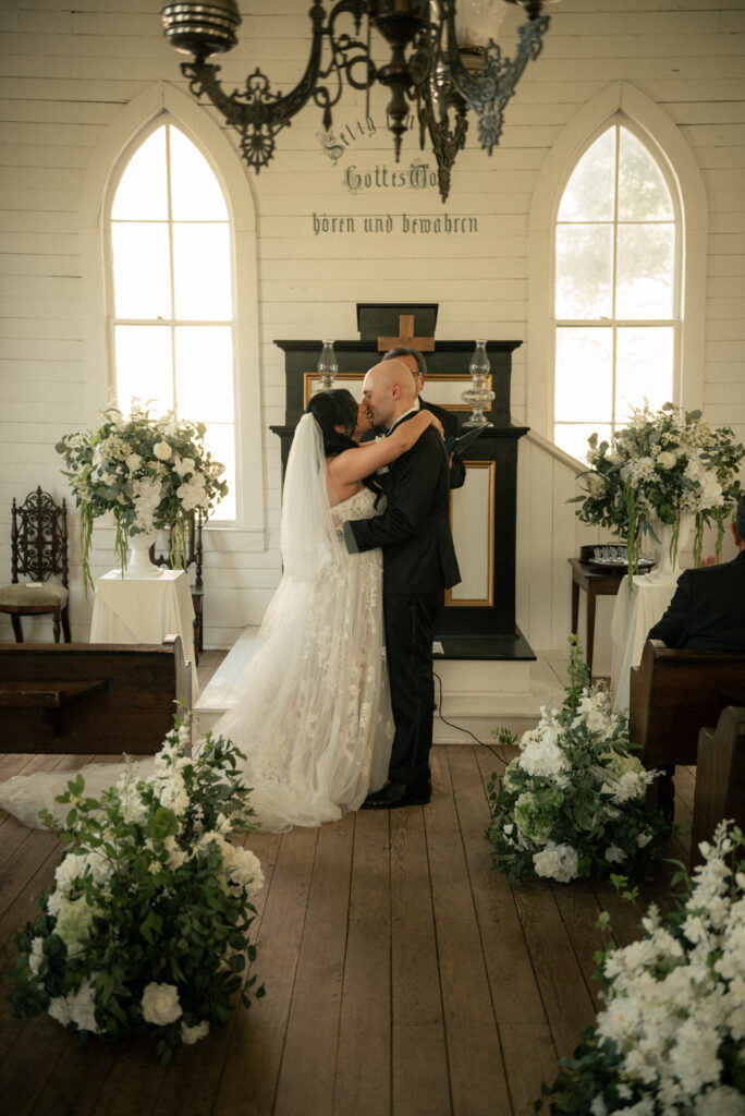First kiss at the end of a chapel wedding ceremony with soft natural light.