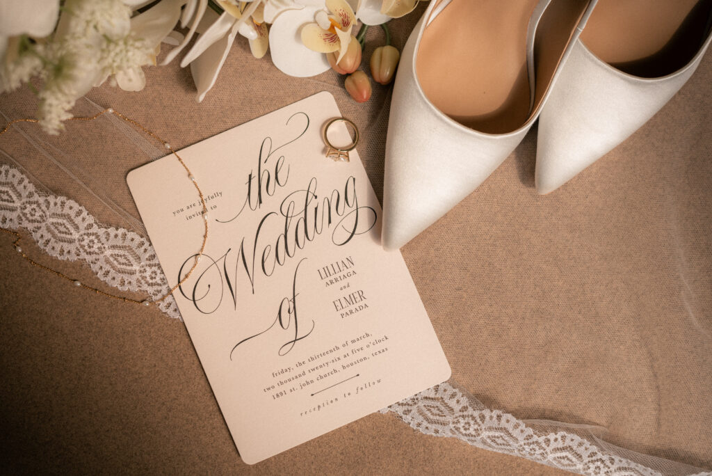 Wedding flat lay with invitation suite, rings, and bridal shoes on neutral fabric.