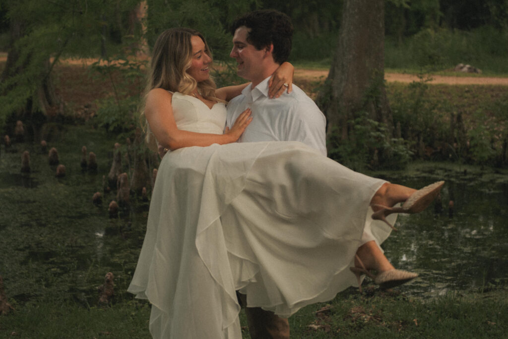 Engaged couple smiling during a playful lift near a pond in a Houston park.